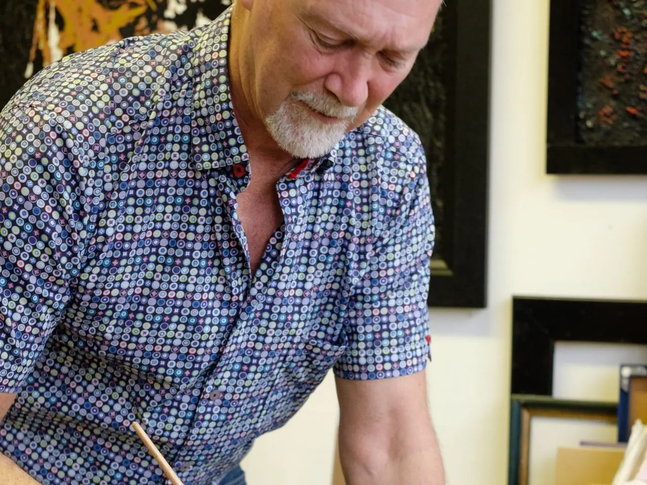 A man in a patterned blue shirt concentrates while painting or working on artwork indoors.