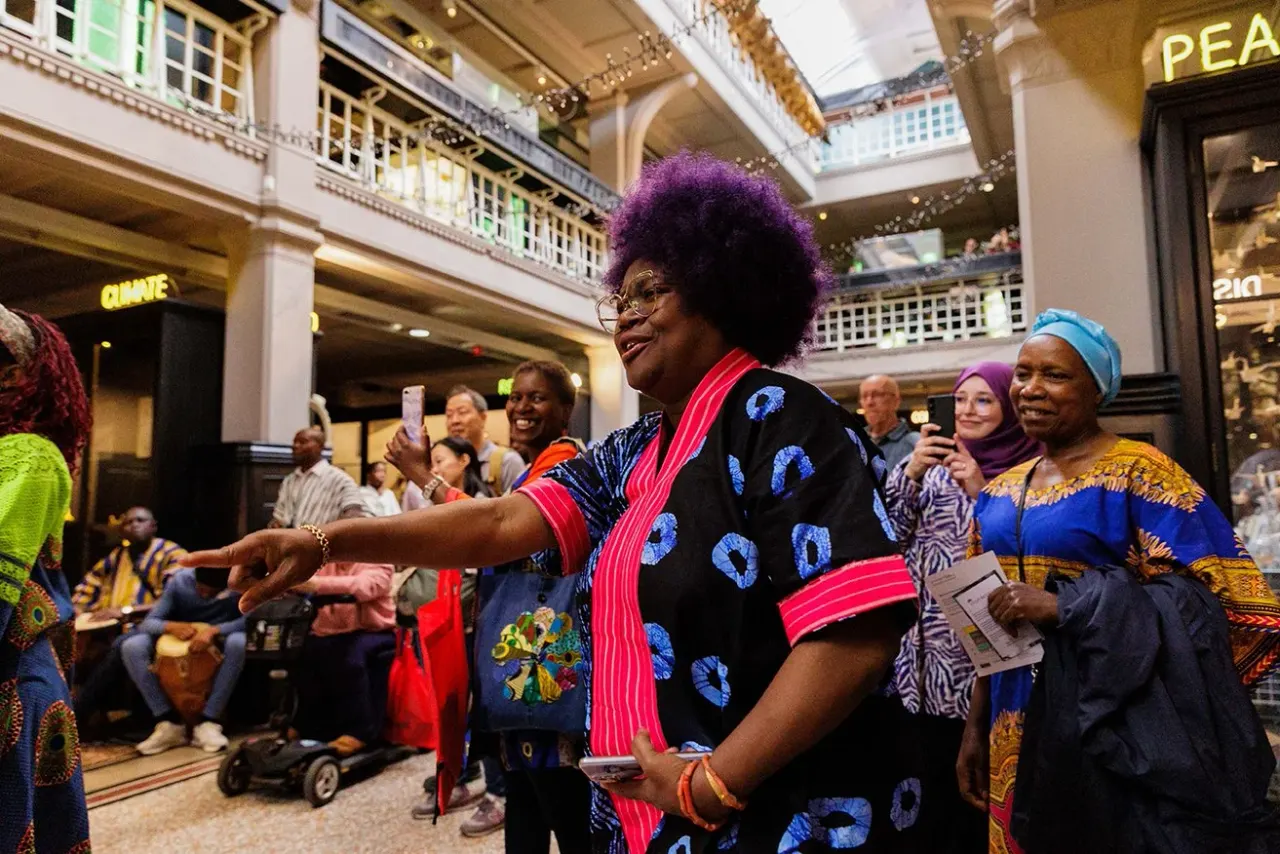 A woman with purple afro hair wearing colourful traditional clothing performs and dances in a multi-level indoor shopping arcade surrounded by spectators.
