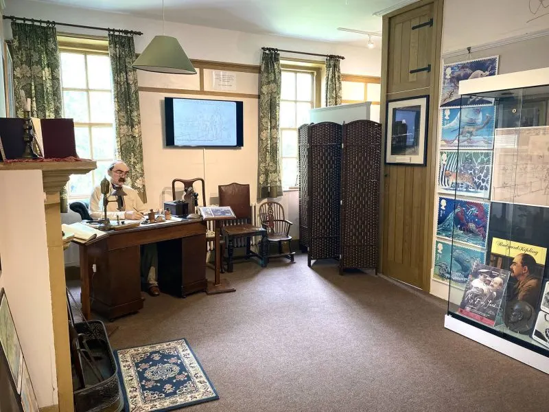 A man sits at a wooden desk in a period room with vintage furnishings, displays and posters on the walls.