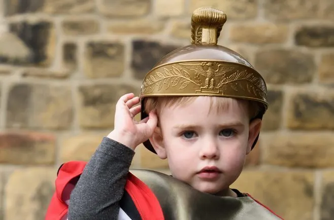 Young child wearing a decorative brass helmet against a stone building backdrop.