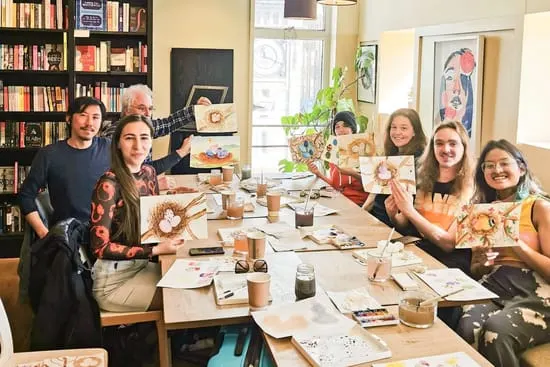 Group of six people seated at a table displaying their painted artwork during an indoor creative workshop session.
