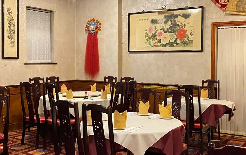 Dining room with round tables set with white tablecloths, folded napkins, and dark wooden chairs, decorated with Asian artwork and a red silk tassel ornament.