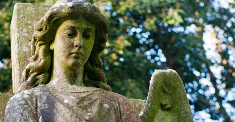 Stone statue of a woman with long wavy hair and downward gaze, covered in moss, amongst trees.