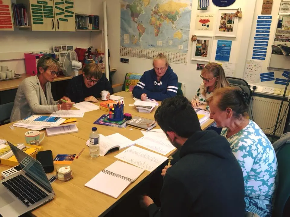 Six adults sit around a wooden table in a classroom, working together on documents and notebooks with educational materials and a world map visible on the walls behind them.