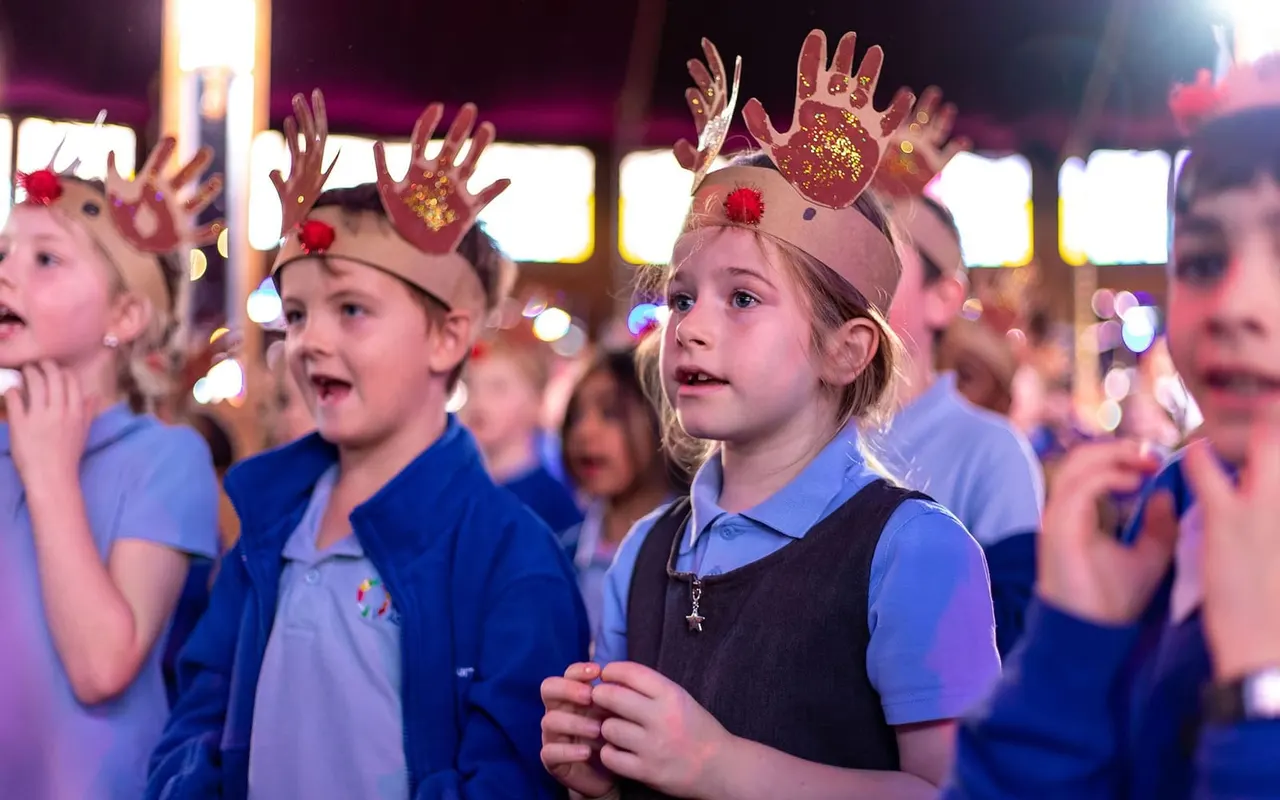 Children wearing blue uniforms and decorated crown headpieces perform on stage with hands raised under coloured lighting.