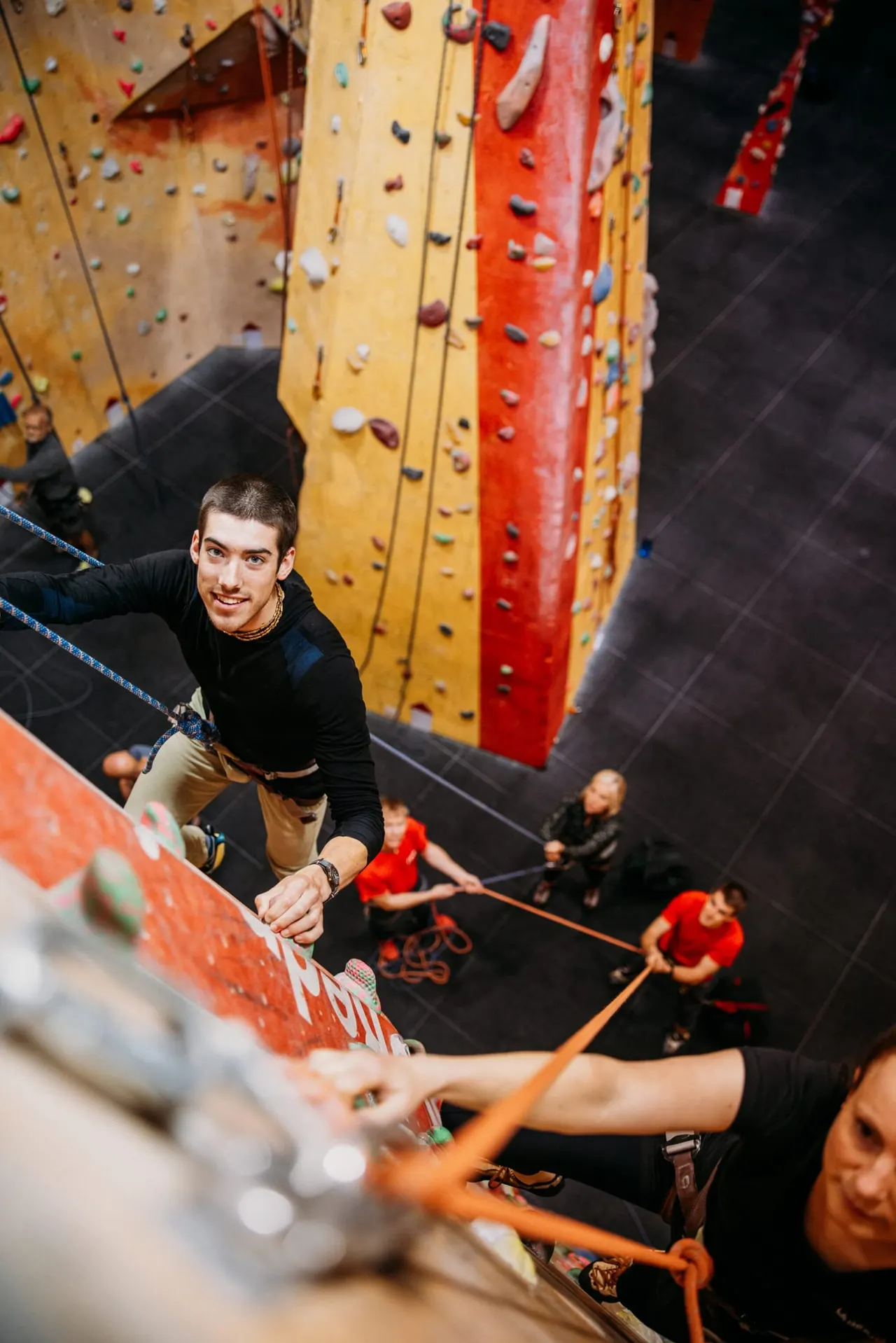 Man indoor rock climbing on a yellow and red wall whilst belayed by others at a climbing centre.