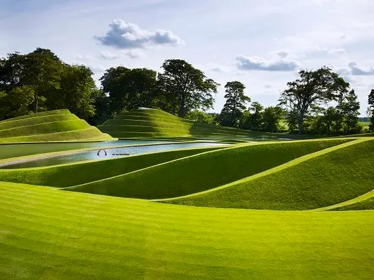 Rolling green lawns with sculptural earthworks and a small water feature in a contemporary art park.