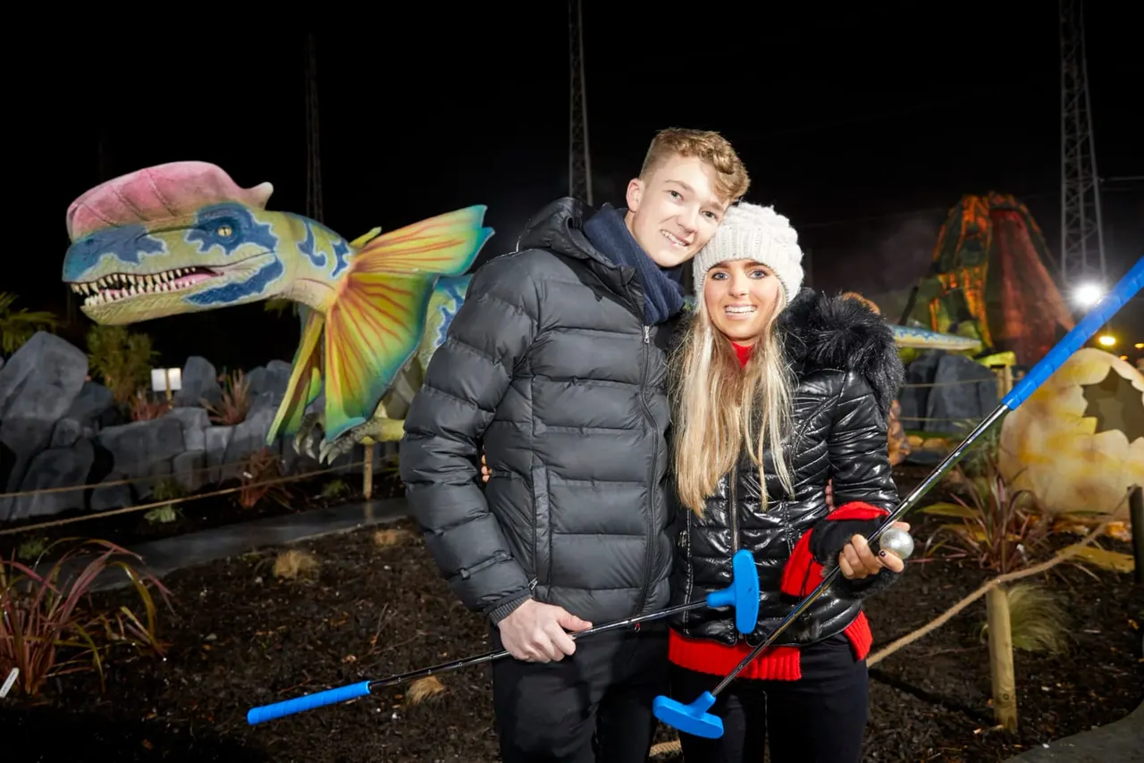 A woman and boy smile at the camera whilst holding putters on a night-time dinosaur-themed mini golf course.