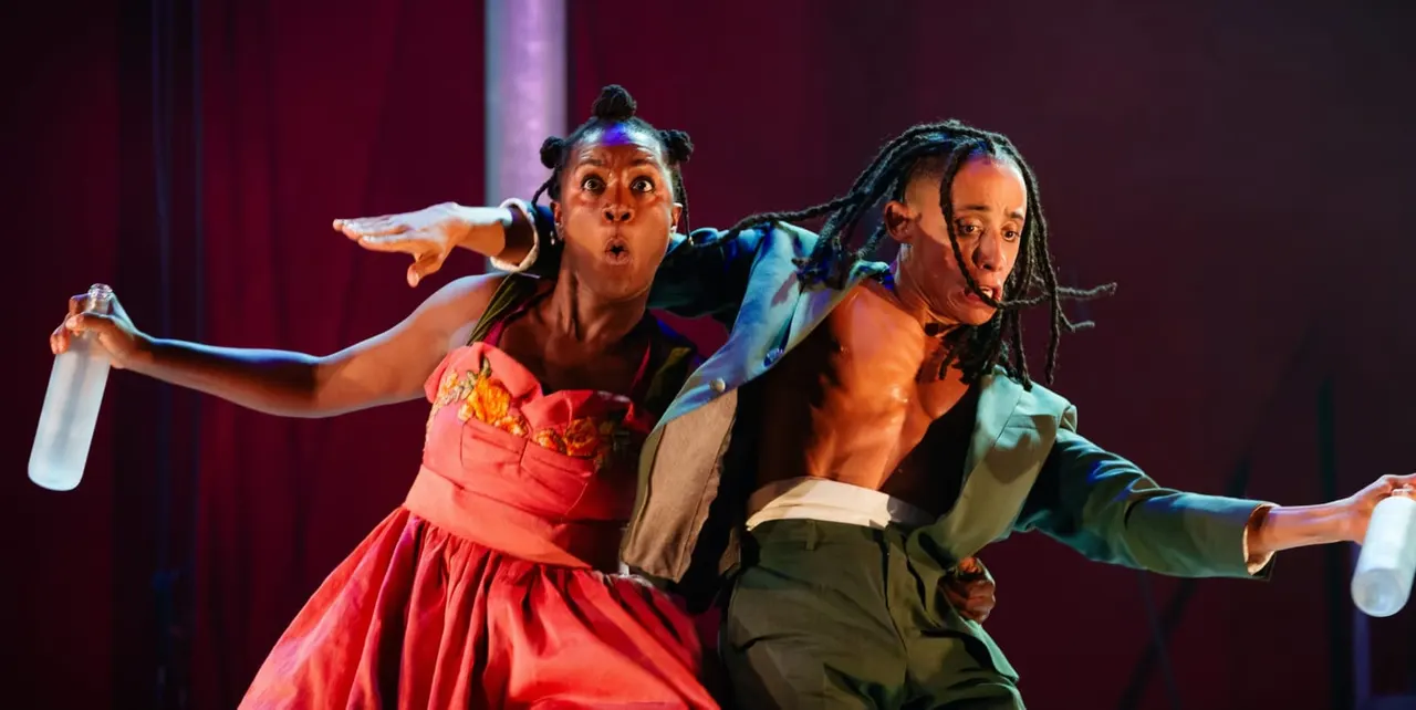 Two performers in colourful costumes strike exaggerated poses on stage under purple lighting during a theatrical production.