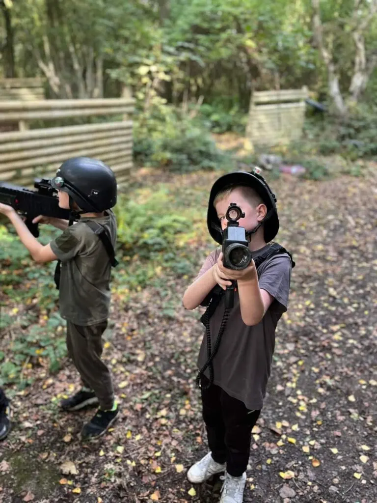 Two children in protective gear and helmets aim paintball guns in a woodland arena with wooden barriers.