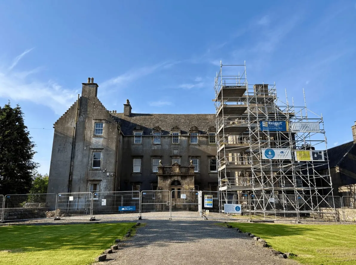 Historic stone mansion with scaffolding on one wing, surrounded by manicured grounds and security fencing under blue sky.