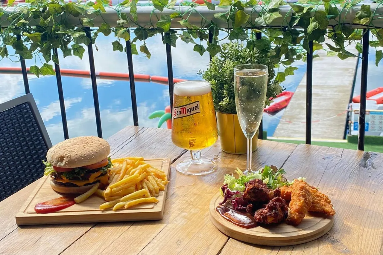 Outdoor waterfront dining table with burger, fries, fried chicken and drinks overlooking a blue lake and water sports equipment.