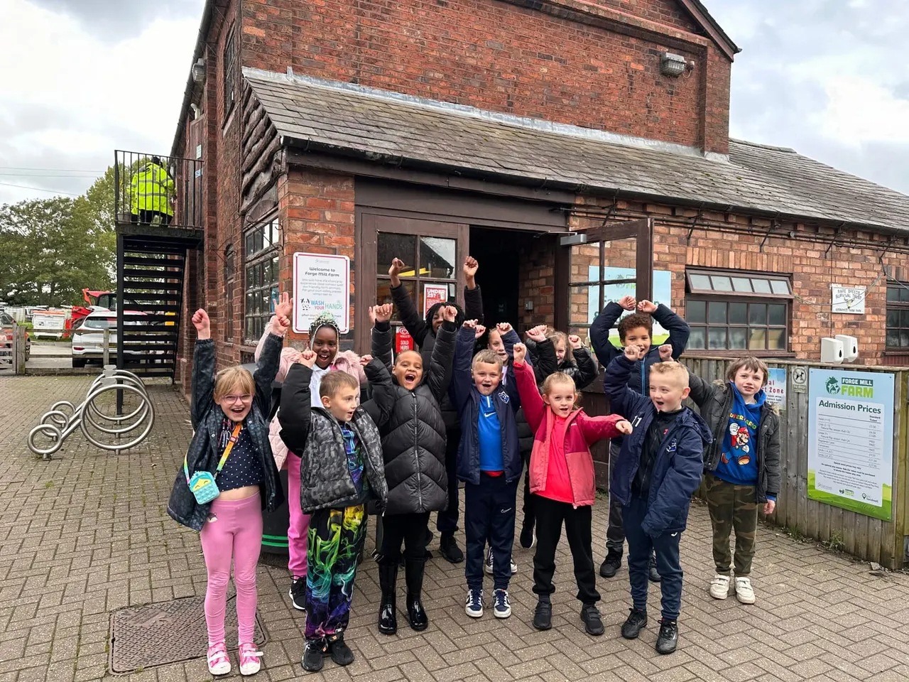 Group of children with arms raised celebrating outside a red brick farm building.