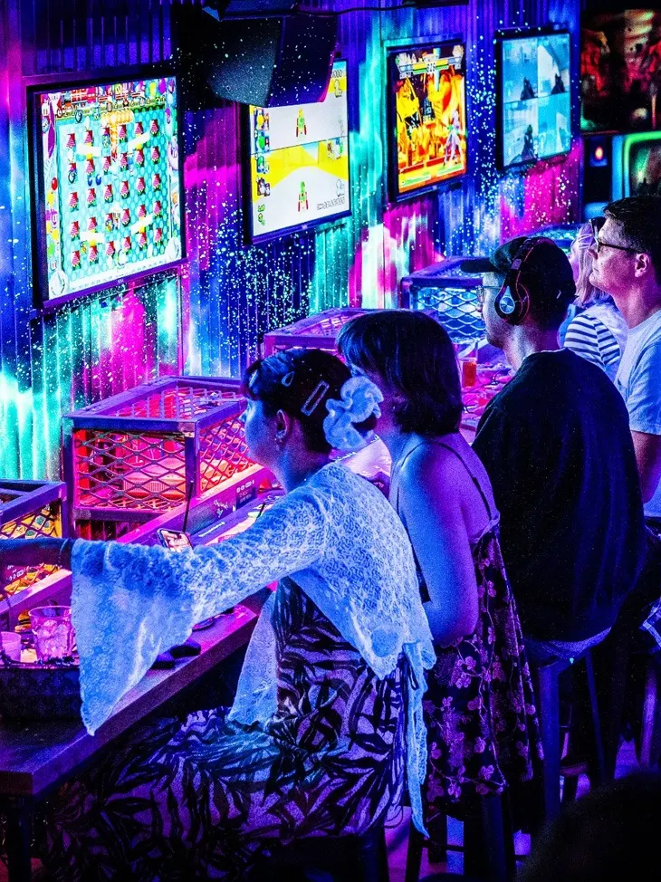 Group of visitors playing arcade games in a neon-lit gaming venue with colourful illuminated screens and gaming stations.