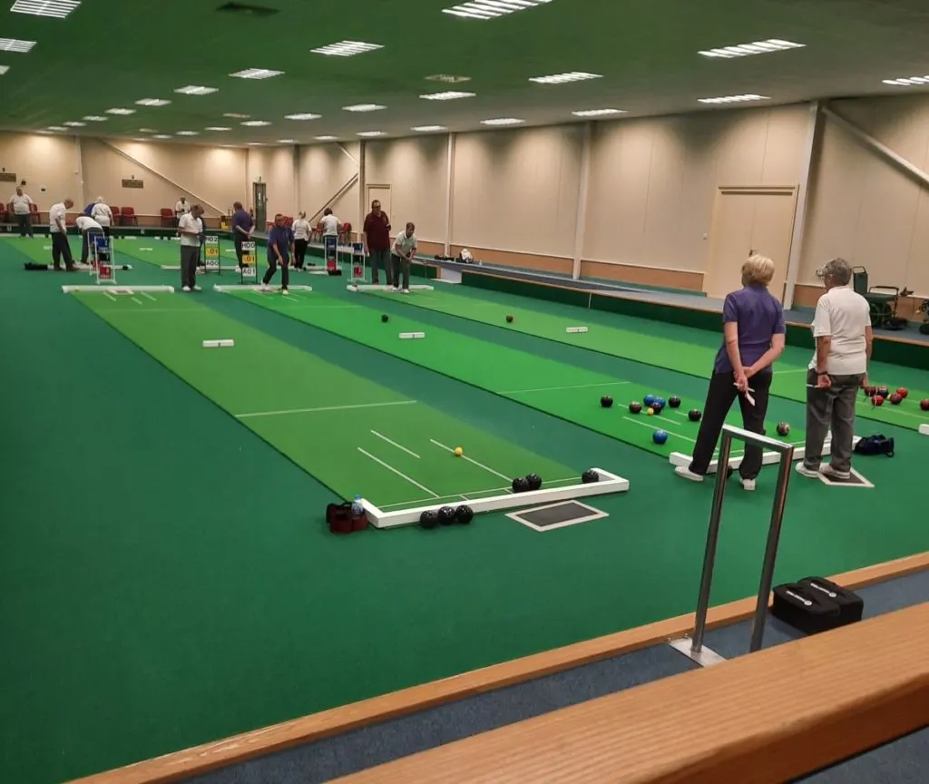 Multiple players practising indoor bowls on several green lanes in a sports hall.