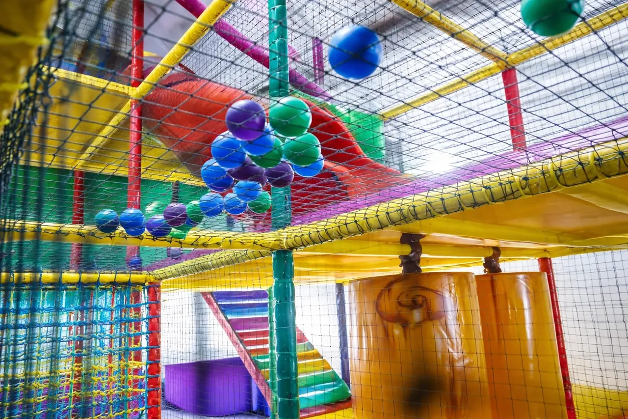 Interior of a colourful children's soft play centre with netting, hanging balls, slides and climbing structures in bright primary colours.