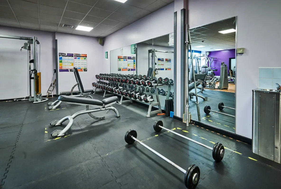 Interior of a gym with dumbbells on racks, barbells on the floor, exercise machines and mirrored walls.