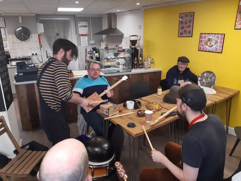 A group of men playing musical instruments together in a bright yellow café space with a counter and kitchen visible in the background.