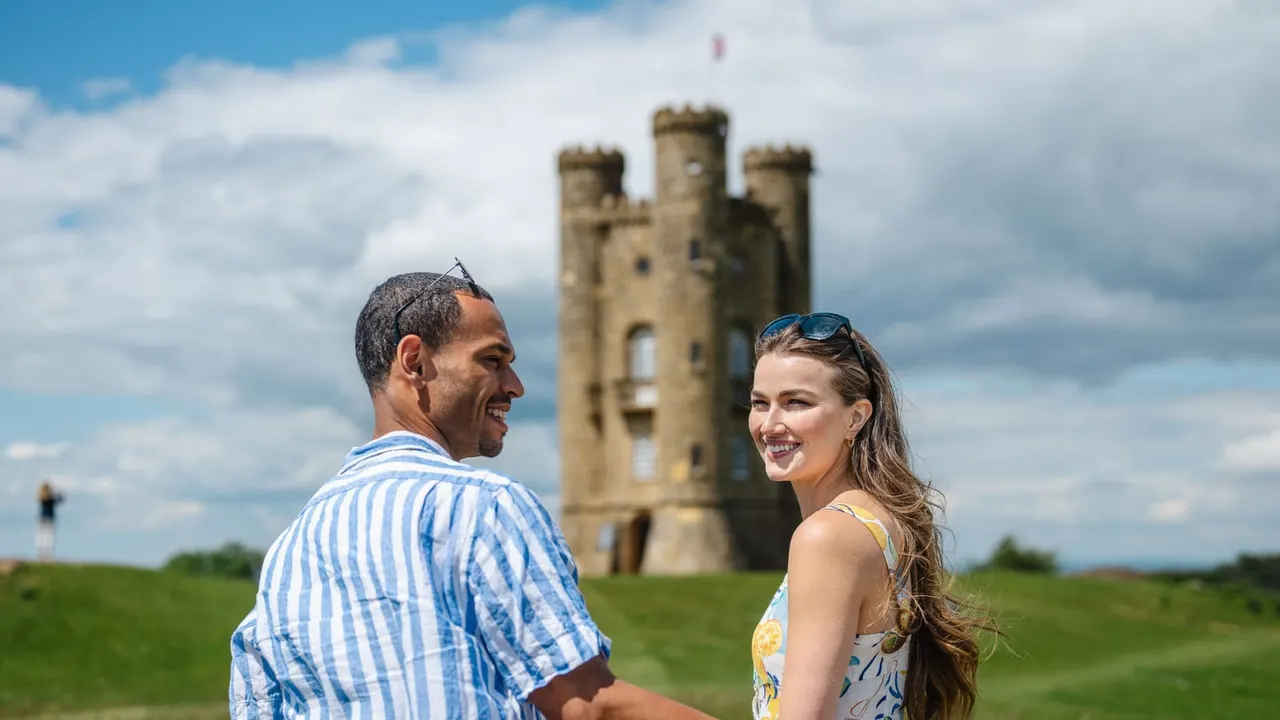 Two visitors smile while standing in a grassy field with a historic stone tower in the background under a partly cloudy sky.