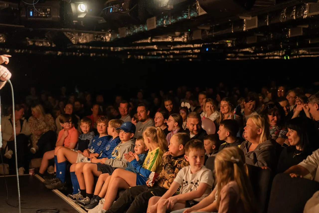 A large audience of adults and children seated on the ground watching a performance on an indoor stage lit by overhead lights.