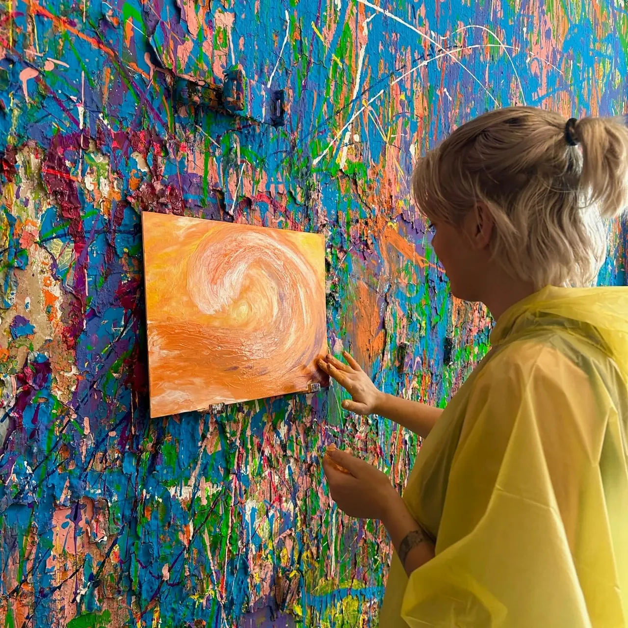 Child in yellow shirt placing orange painting on vibrant multicoloured splattered wall display.