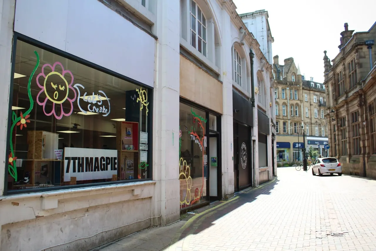 A colourful storefront with painted flower designs on its window, situated on a sunny pedestrian street lined with historic stone buildings.