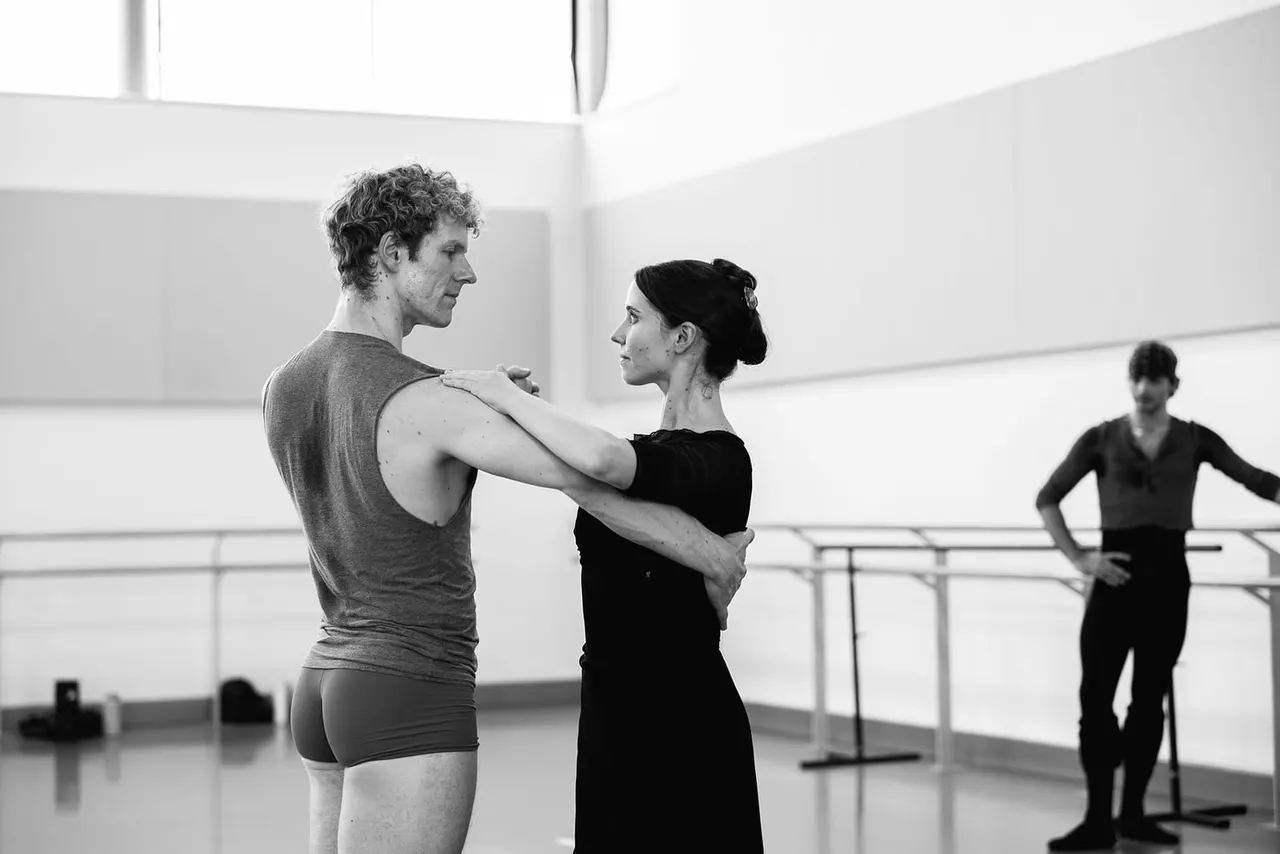 A dance instructor guides a male dancer during a rehearsal in a bright studio with a barre, whilst another dancer watches in the background.