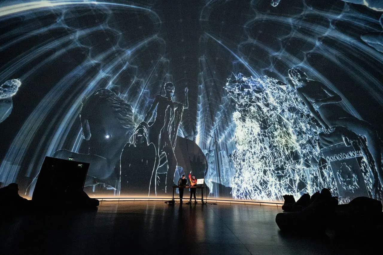 Two performers stand on stage beneath a massive immersive projection of architectural and figurative imagery in blue and white tones.