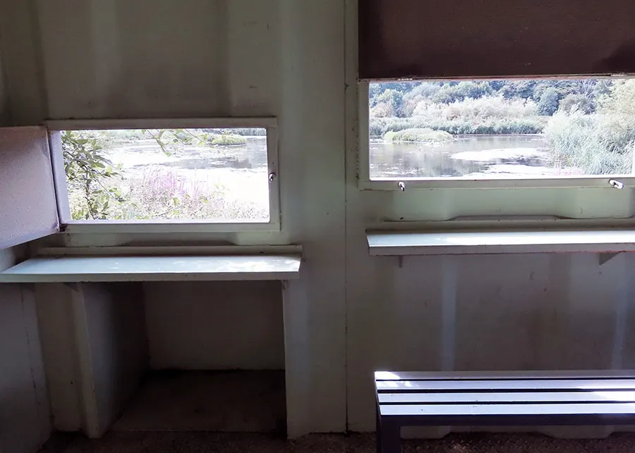 Interior of a viewing hide with benches and windows overlooking a river and wooded landscape.