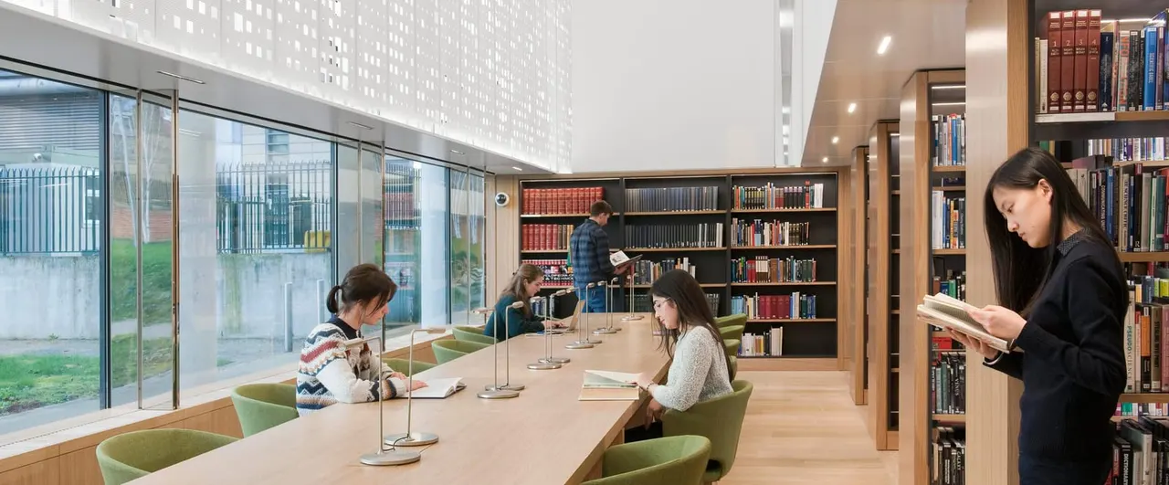 Modern library interior with visitors studying and browsing books at tables and shelves, with natural light from large windows.