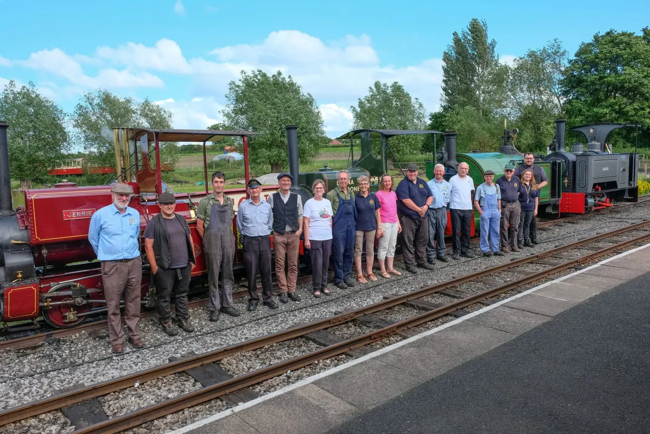 A group of railway volunteers and staff stand alongside heritage steam locomotives on the narrow-gauge track on a sunny day.