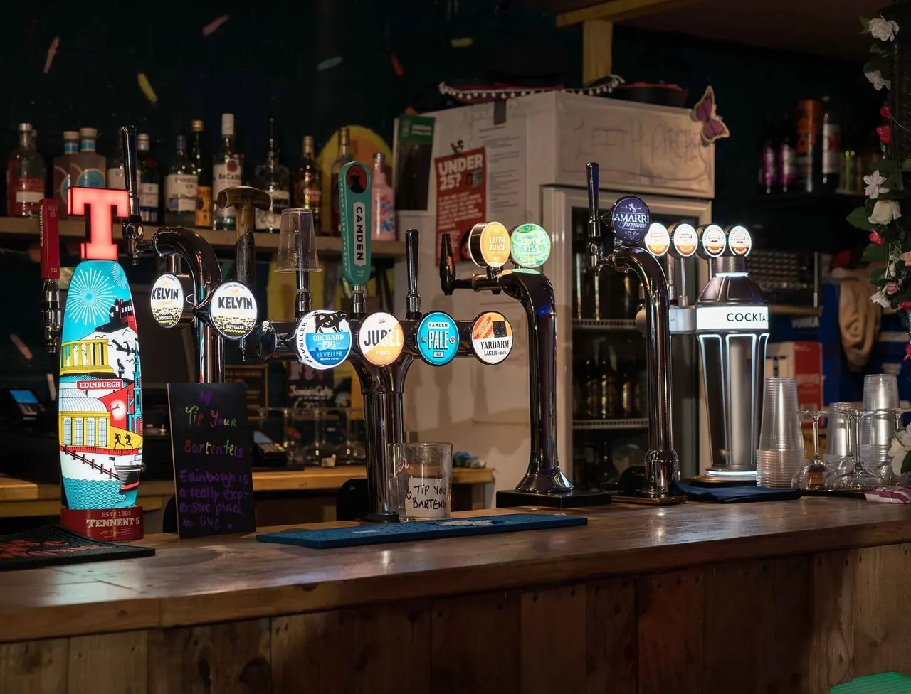 Bar counter displaying multiple beer taps with branded pump clips, bottles on shelves behind, and glassware in a dimly lit pub setting.