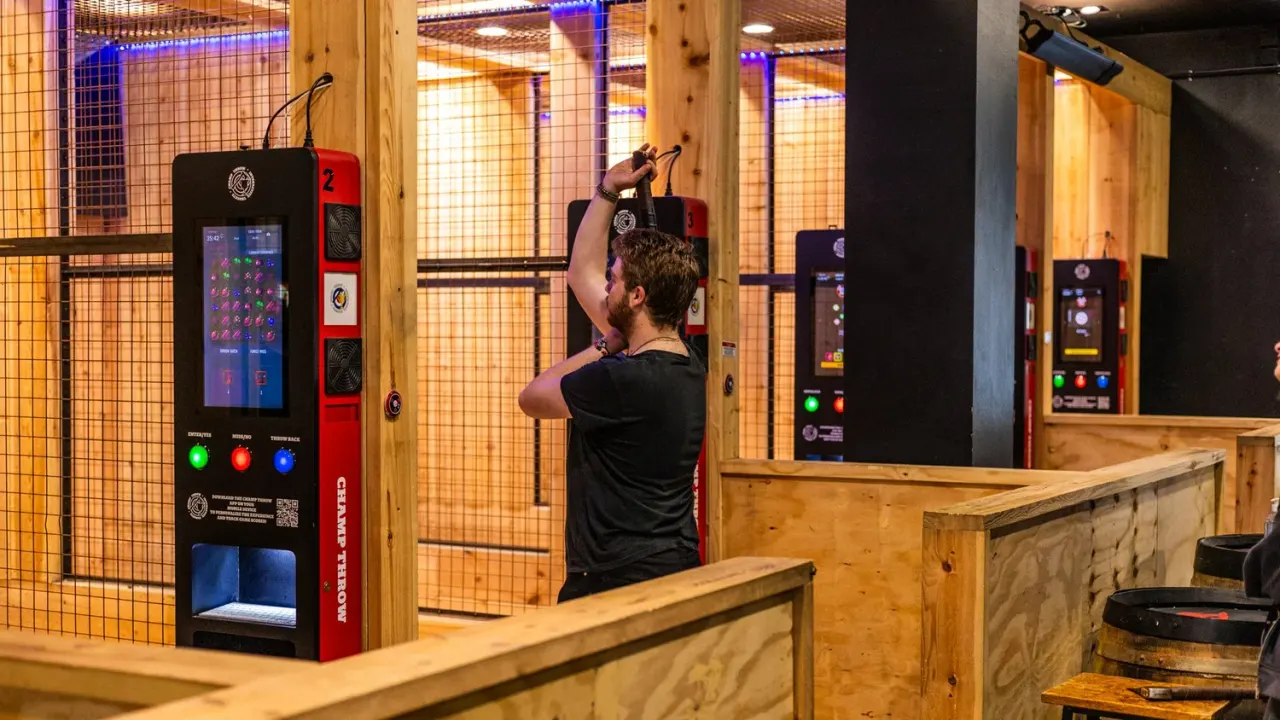 Man throwing axe at target in indoor throwing range with wooden booths and electronic scoring machines.