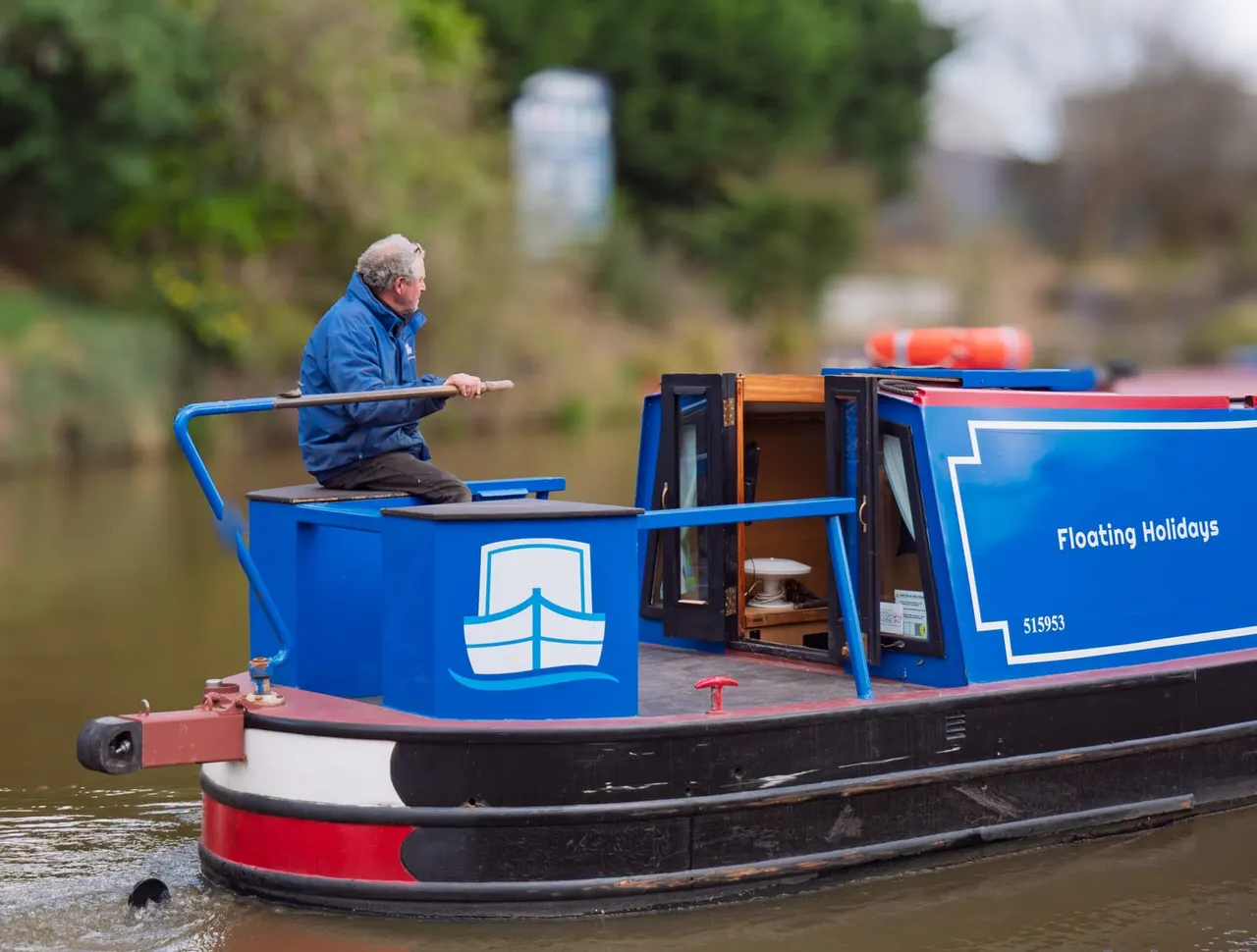 An older man in a blue jacket steers a blue and red narrowboat along a calm waterway surrounded by green vegetation.