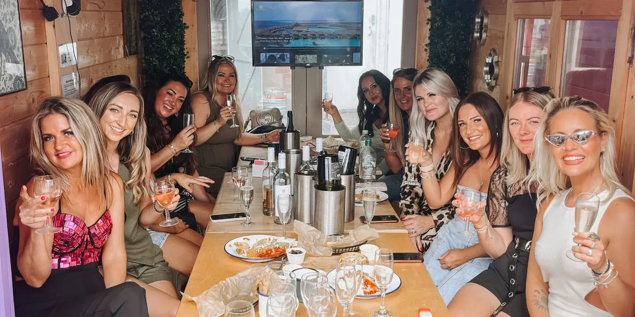 A group of women holding drinks at a dining table at an indoor venue, smiling at the camera during a celebration.