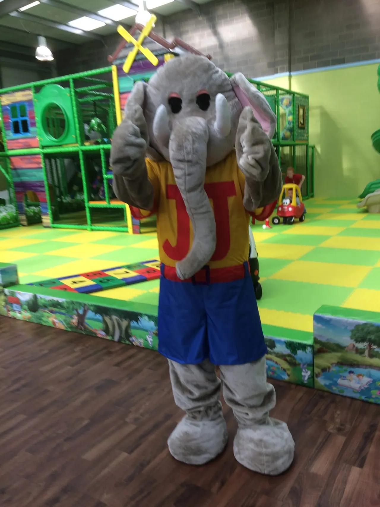 A mascot in an elephant costume stands in a colourful soft play facility with padded flooring and climbing structures.