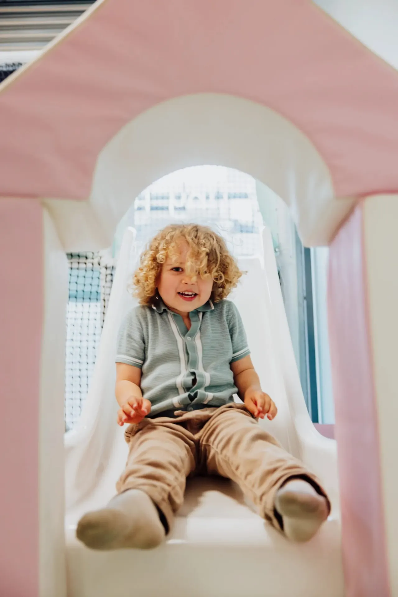 A young child with curly hair slides down a pink and white play tunnel, smiling.