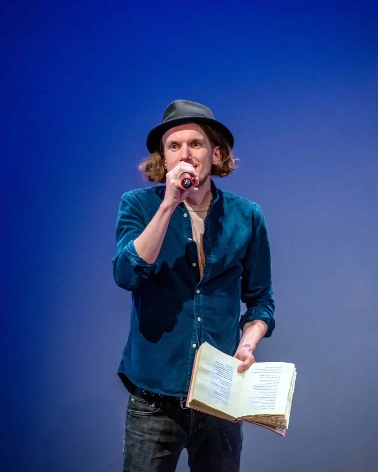 A performer on stage holding a microphone and an open book, wearing a denim shirt and hat against a blue backdrop.