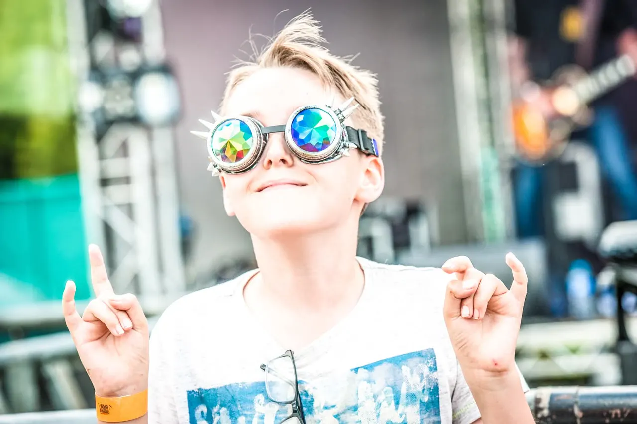 Child wearing rainbow kaleidoscope goggles at an outdoor event, gesturing with both hands towards the camera.