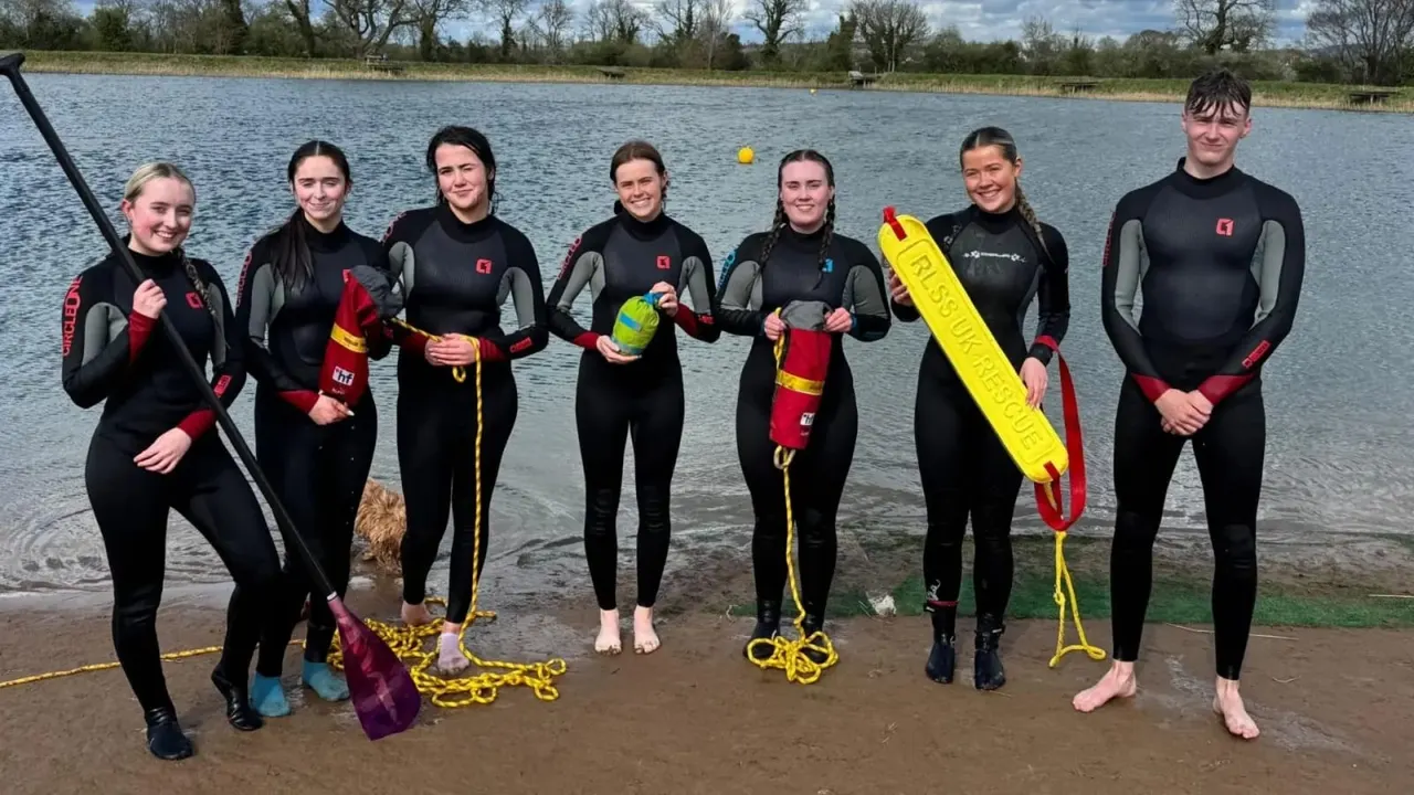 Group of seven people in wetsuits standing on a beach holding watersports equipment with a lake behind them.