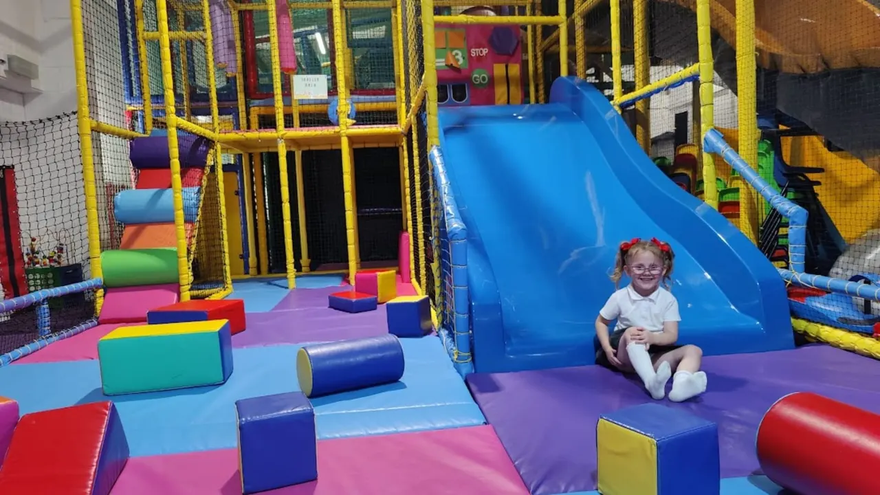 Young child sitting at the bottom of a large blue slide in a colourful indoor soft play area.