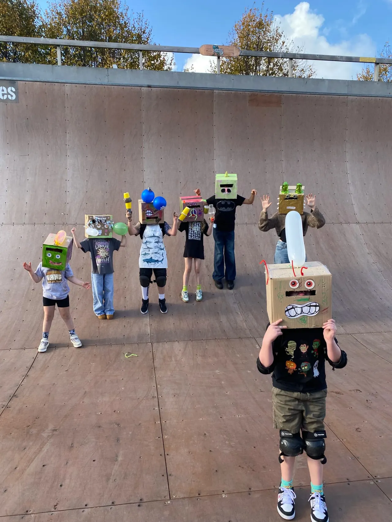 Group of children wearing cardboard box monster masks and costumes posed in a skate bowl against a concrete wall.
