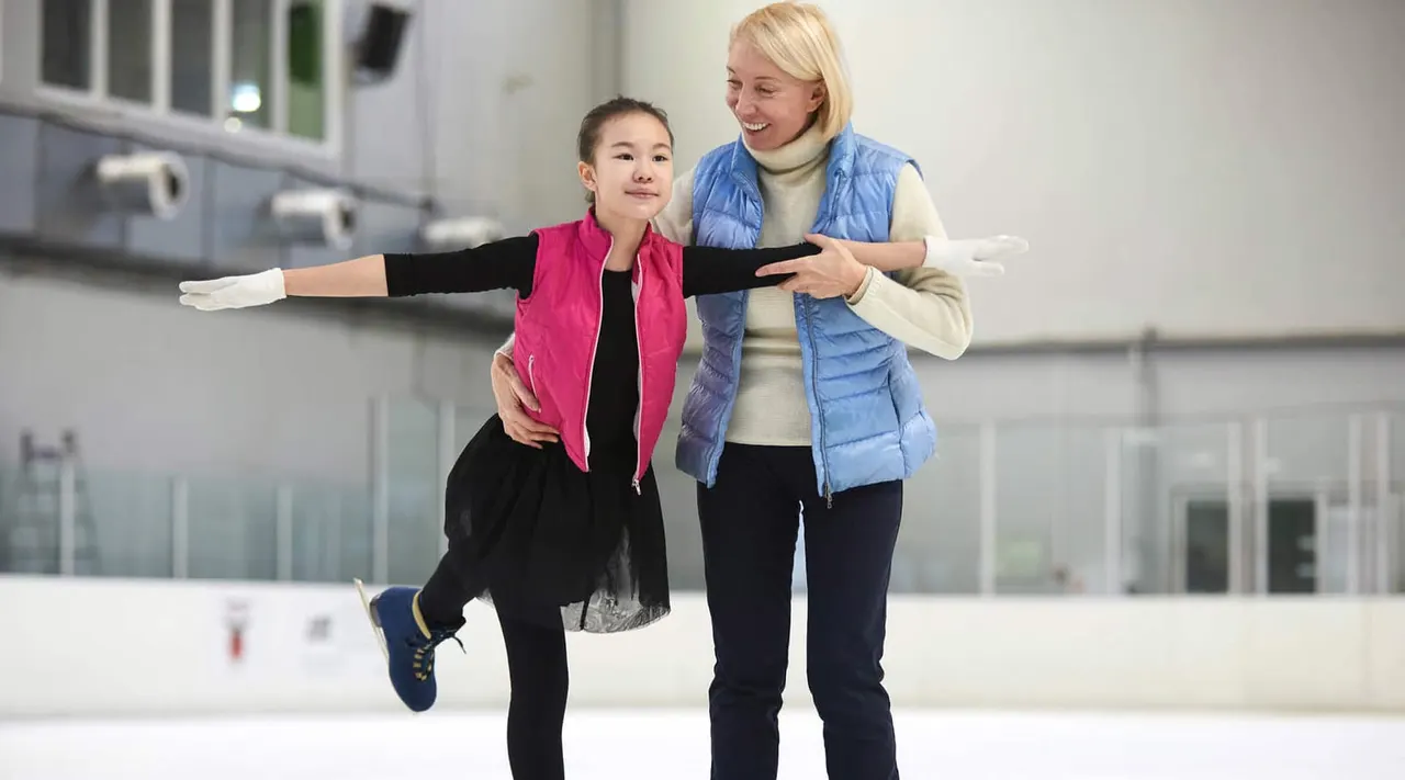 A young girl ice skating with arms outstretched whilst an adult instructor guides her on an indoor ice rink.