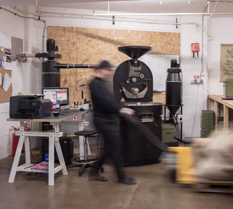 Person working at a coffee roastery with large roasting equipment and industrial machinery.