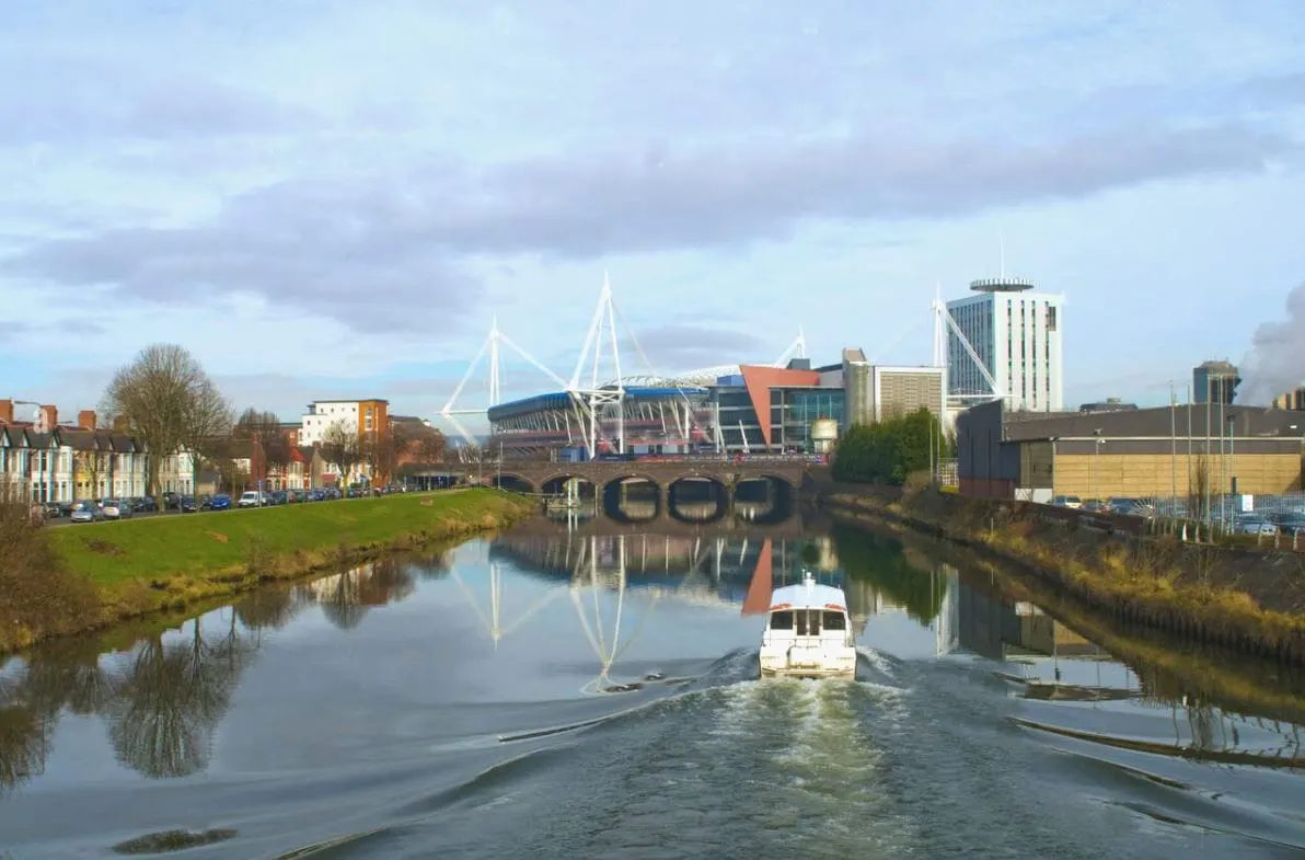 A white boat travels along a canal towards a large modern stadium and cable-stayed bridge, with city buildings and bare trees lining the waterway.