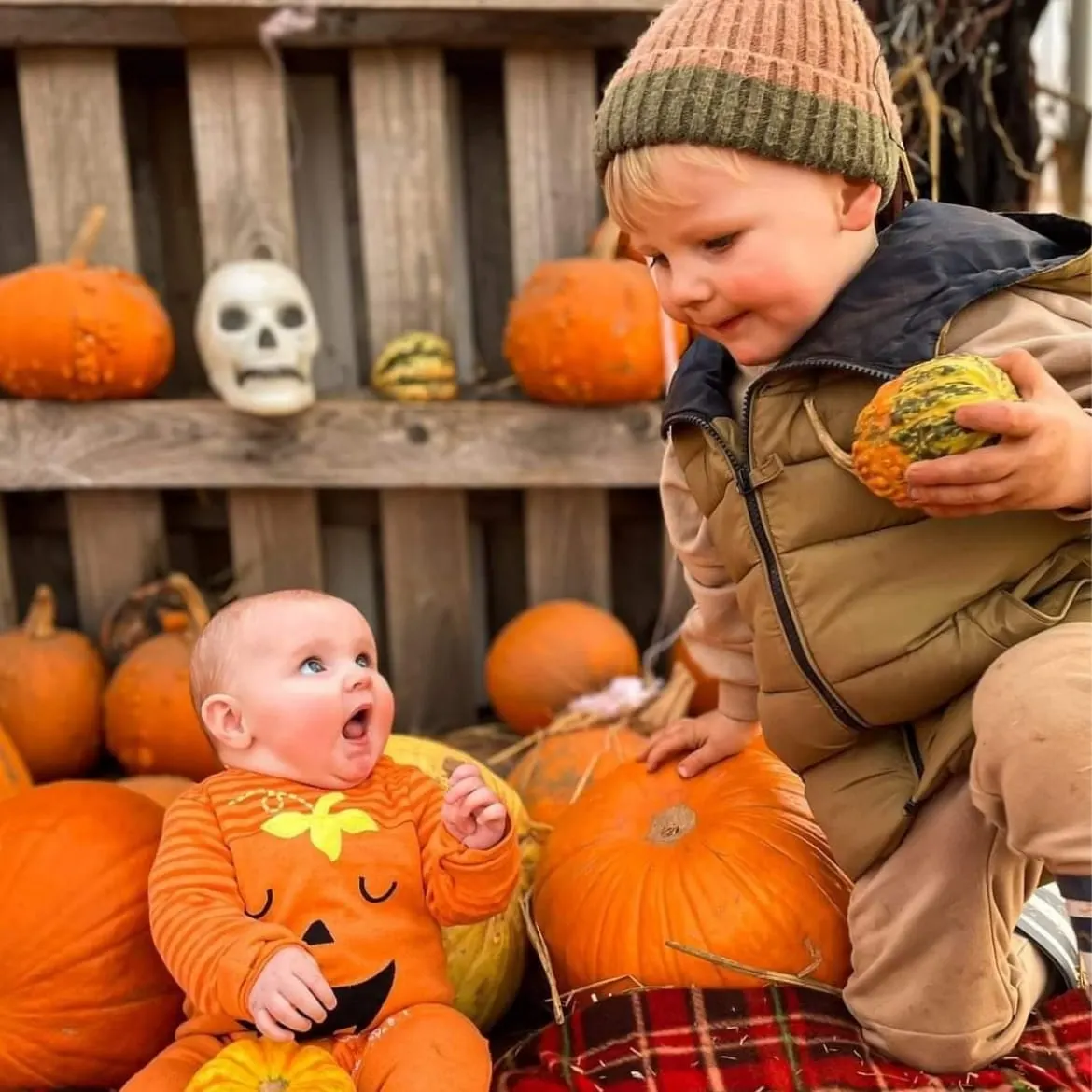 Young child in striped pumpkin outfit sits among pumpkins whilst older child in knitted hat holds a small gourd nearby.