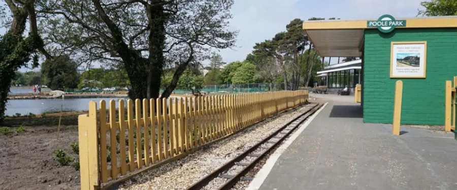 Narrow-gauge railway track alongside yellow fencing with a green station building and lake visible beyond.