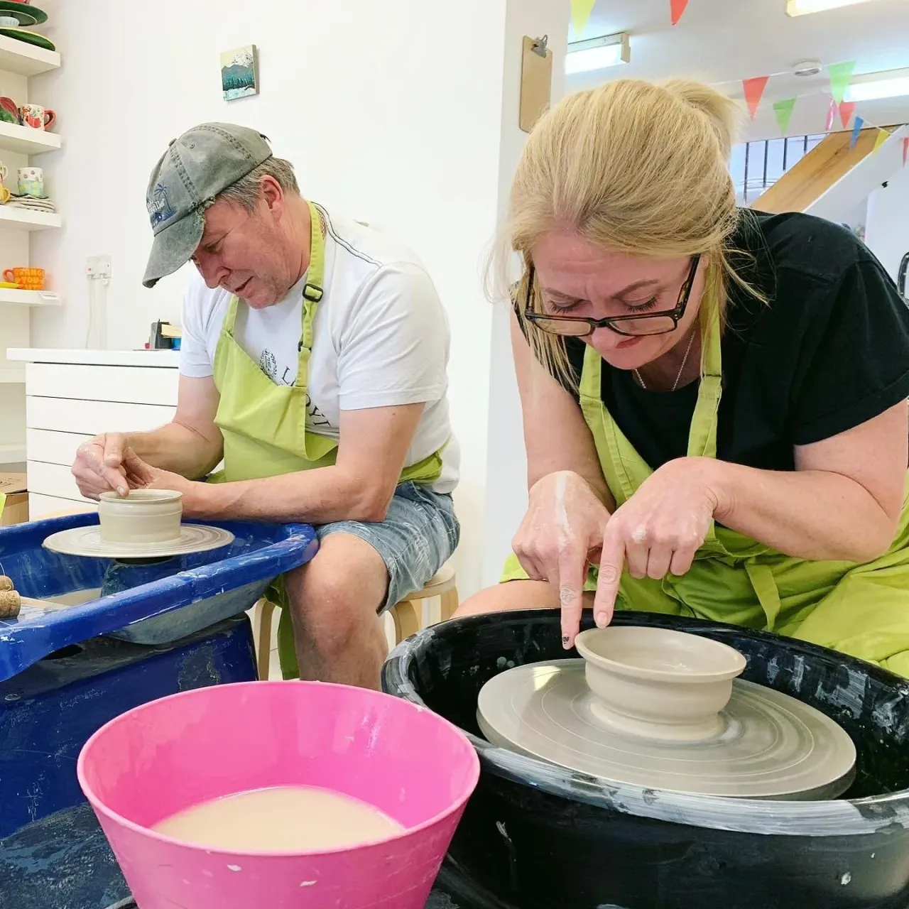 Two adults working at pottery wheels, shaping clay bowls in a bright studio with coloured bunting.