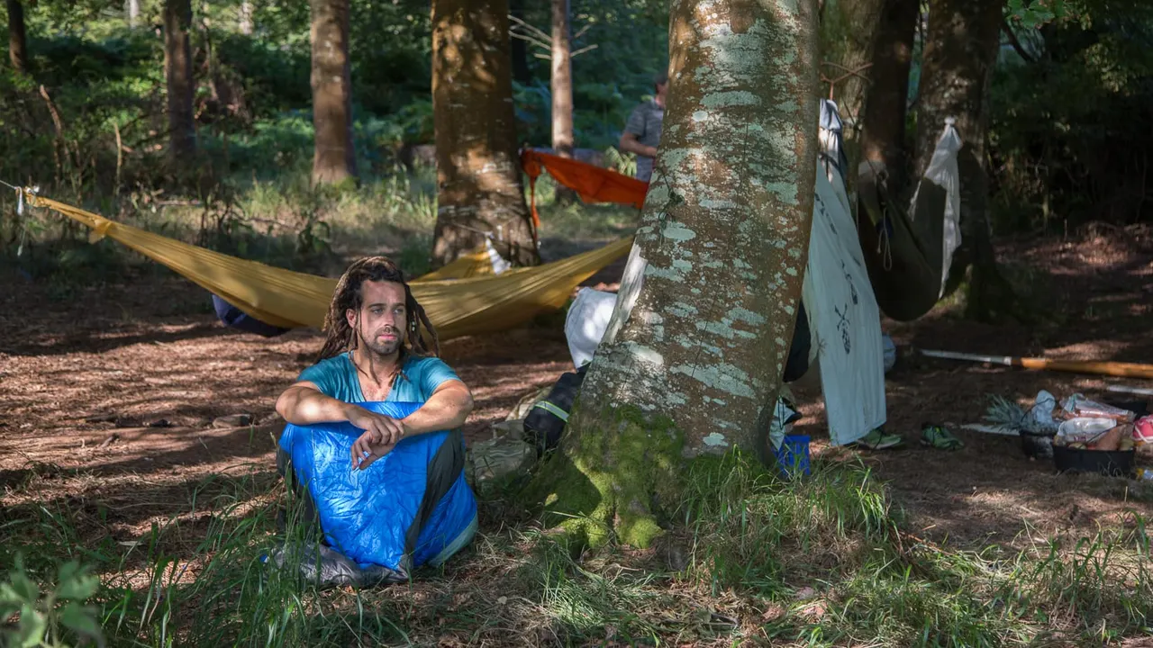 PHOTOGRAPH

Man crouching by a tree in a woodland campsite with hammocks and camping equipment visible.