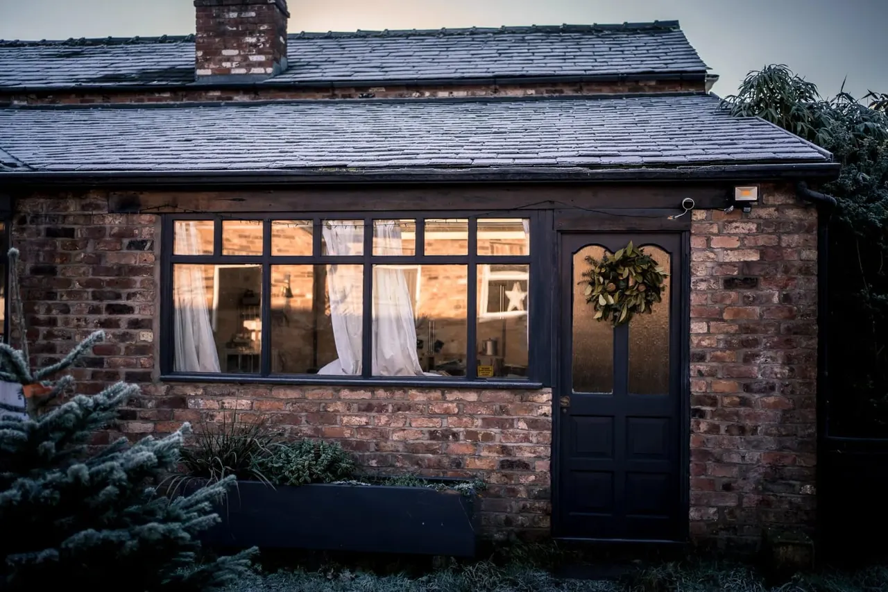 Brick cottage with dark-framed windows and door, illuminated interior, chimney visible, potted plants and greenery outside.